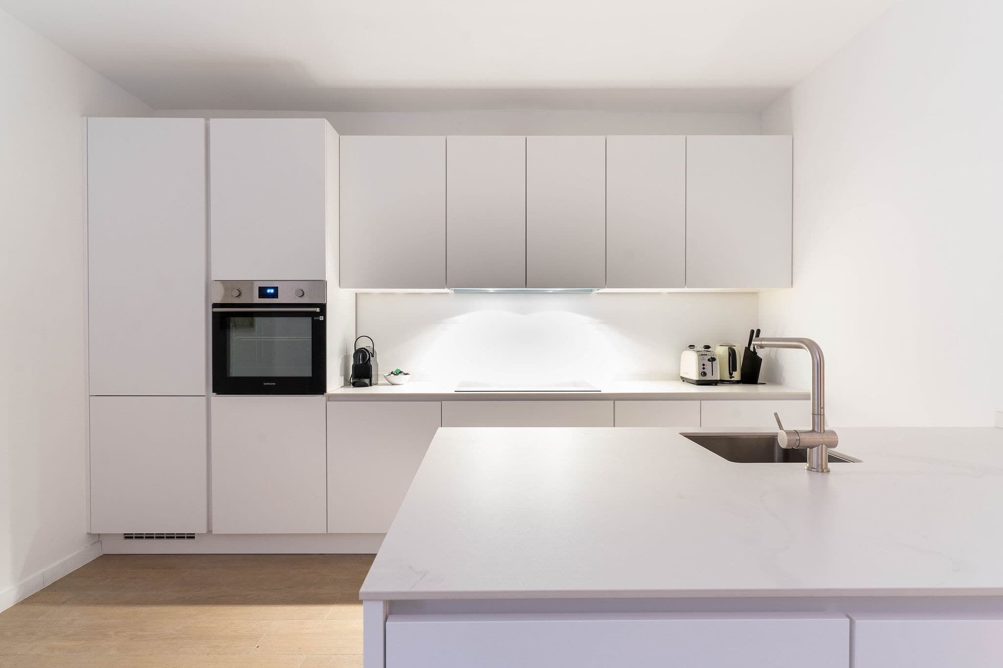 A modern kitchen with white cabinetry, a built-in oven, a stainless steel sink, and a countertop with a toaster.