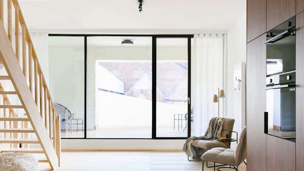 A modern living room with a wooden staircase, a sofa, a dining table, and a large sliding glass door leading to an outdoor area. - Magnificent 3BR home in Antwerp (Antwerpen)