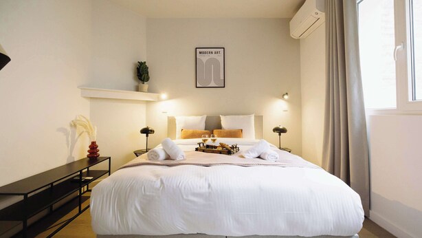 Ein modernes Schlafzimmer mit einem großen Bett, Nachttischlampen, einer Wand-Klimaanlage und einem gerahmten Kunstwerk.