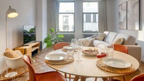 En moderne stue med spisebord dækket til to personer, sofa, fjernsyn og store vinduer med gardiner.