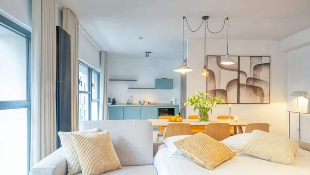 A modern hotel room with a sofa, a dining table, and a kitchen area in the background.