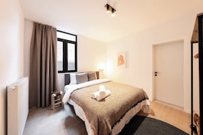 Ein Hotelzimmer mit Bett, Fenster mit Vorhängen, Heizkörper, kleinem Tisch und Tür.