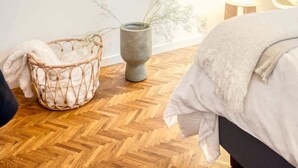 A bedroom with a wooden herringbone floor, a wicker basket, a grey vase with dried flowers, a bedside lamp, and a bed with a white blanket. - Bright 2BR Flat in Antwerp City (Antwerpen)