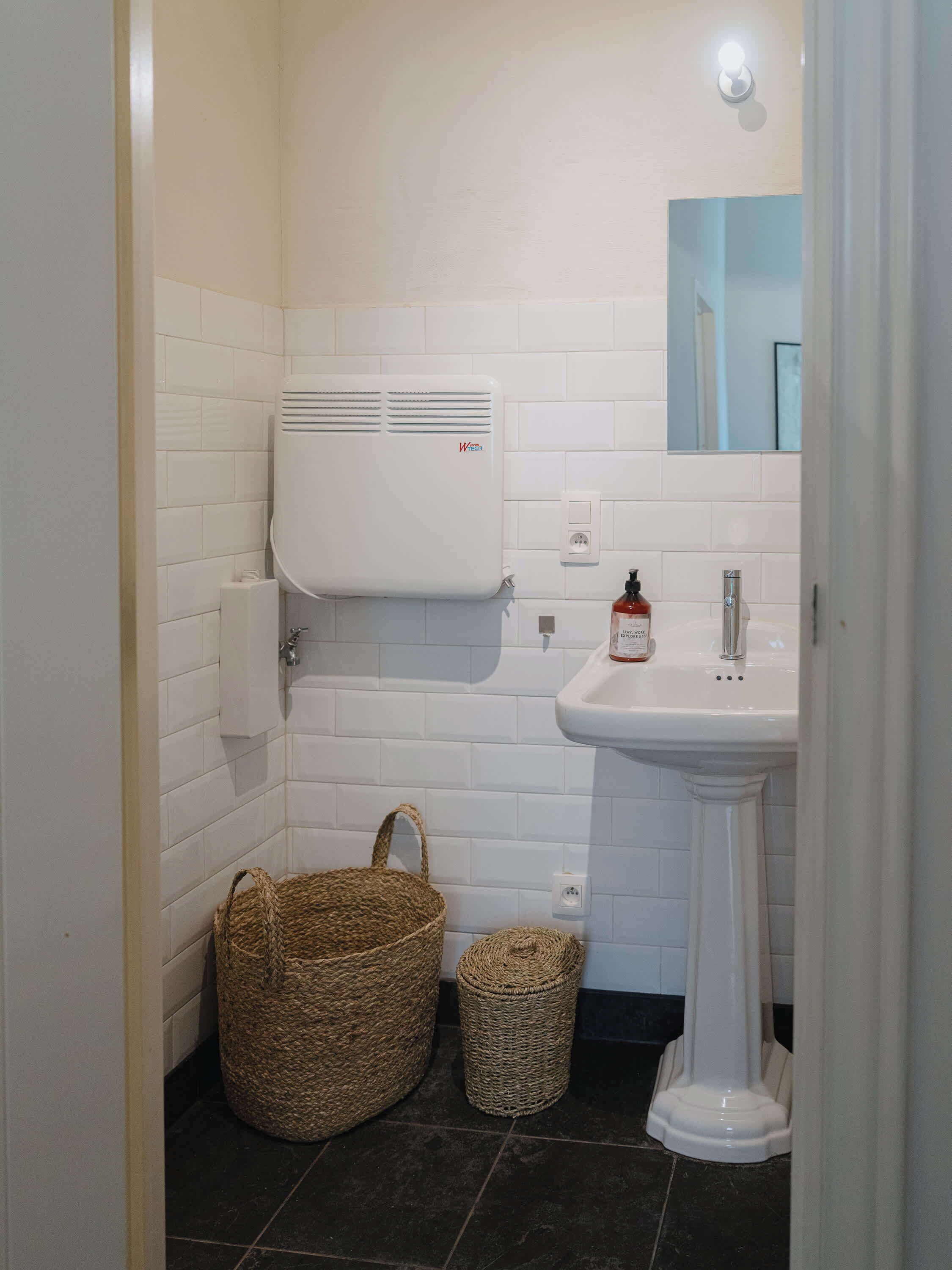 Een badkamer met witte betegelde wanden, een wit wastafeltje, een handdroger, een geweven mand en een kleine prullenbak.