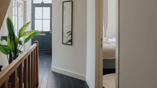 A hallway with a wooden railing, a potted plant, a framed picture, and a bed with a white blanket in the adjacent room. - Spacious Triplex Apartment in Mechelen (Mechelen)