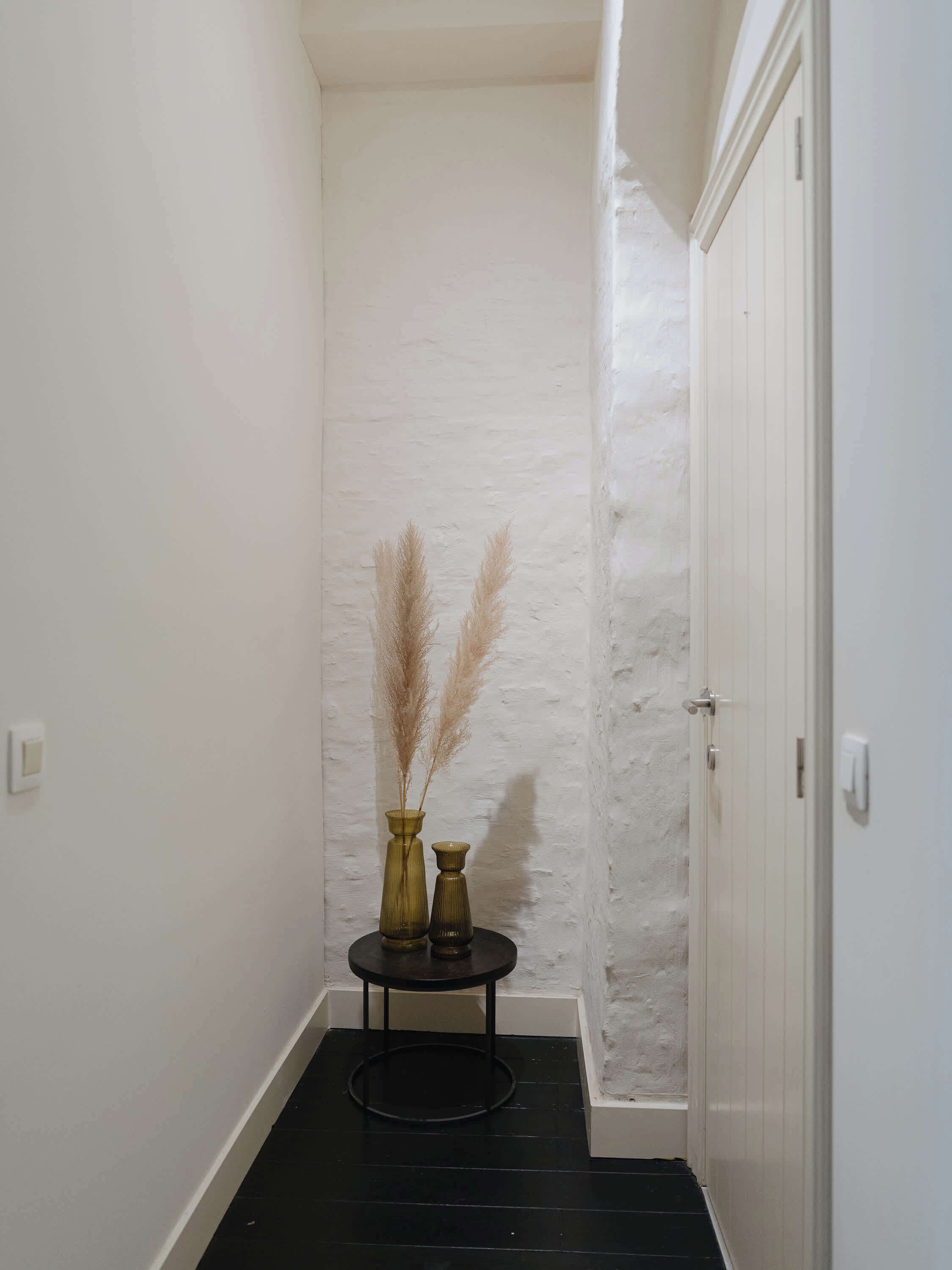 A narrow hallway with white walls and a black floor. There is a small round table with two vases containing pampas grass.