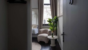 Et hotelværelse med en seng, en stol, et lille bord med en plante og en flaske, samt et vindue med gardiner.
