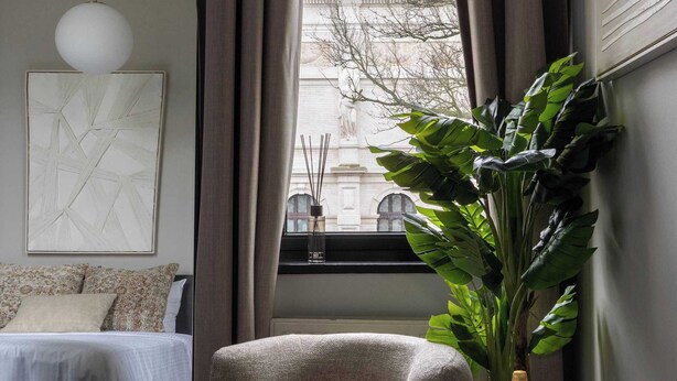 Et moderne soveværelse med en seng, et rundt bord med plante og vinglas, samt udsigt over byen fra et vindue.