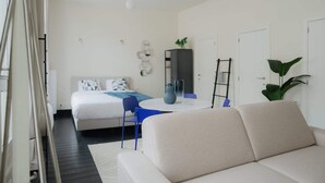 A modern hotel room with a bed, a sofa, a dining table, and a plant.