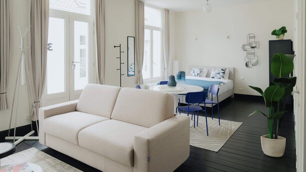 A modern hotel room with a sofa, a bed, a dining area, and a large window.