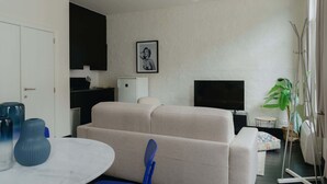 A living room with a sofa, a TV, a dining table with chairs, a small kitchen area, and a plant.