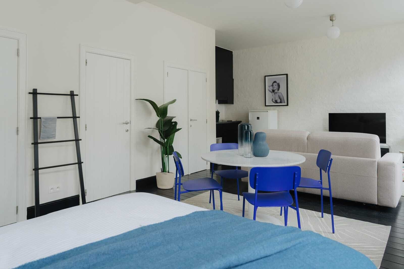 A modern hotel room with a bed, a round table with chairs, a sofa, a potted plant, and a ladder against the wall.