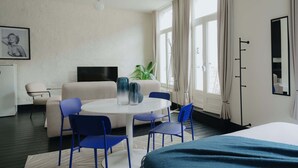 A modern hotel room with a bed, a round dining table with chairs, a sofa, a TV, and a plant.