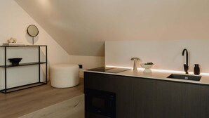 A modern kitchen with a dark countertop, a built-in oven, a round mirror, and a small round table with a bowl of fruit. - Elegant and Spacious Suites (Antwerpen)