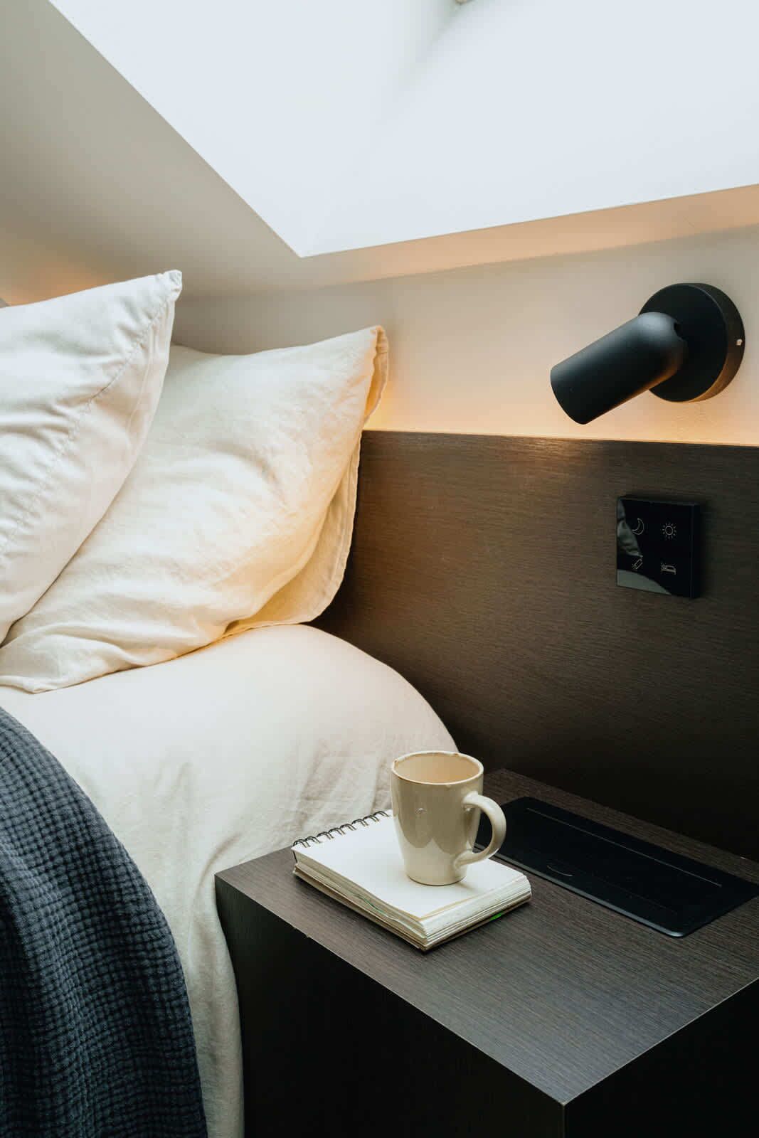 Een net opgemaakt bed met een kussen, een nachtkastje met een notitieboekje en een mok, en een wandlamp.