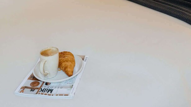 A hotel room with a bed, a bedside table with a lamp, a framed artwork on the wall, and a breakfast tray with a croissant and a cup of coffee. - Elegant and Spacious Suites (Antwerpen)