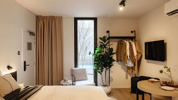 Una moderna camera d'albergo con un letto, un comodino, una piccola sedia, un tavolo da pranzo, una pianta in vaso, un appendiabiti con vestiti e una TV a parete.