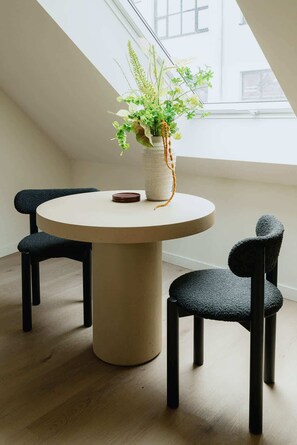 Une table ronde avec un vase de fleurs, deux chaises et une fenêtre dans une pièce dont le sol est recouvert de parquet.