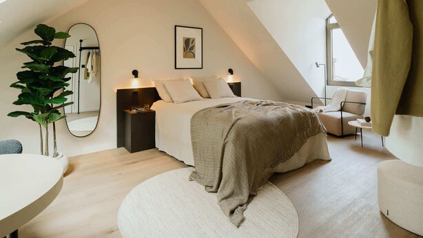 A modern bedroom with a sloped ceiling, a bed with a beige blanket, a bedside table with a lamp, a large mirror, and a potted plant. - Spacious Suite for 4 with Kitchenette (Antwerpen)