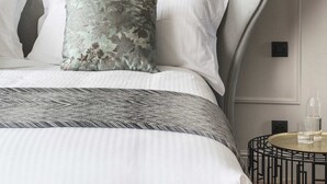 Een net opgemaakt bed met een grijs hoofdeinde, witte kussens en een geborduurde plaid. Aan de zijkant staat een klein rond tafeltje met een glas en een fles.