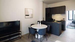 Una sala moderna con un televisor de pantalla plana, una mesa de comedor redonda con sillas, una zona de cocina con armarios oscuros y un gran ventanal que proporciona luz natural.