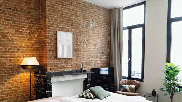 A modern bedroom with a brick wall, a large bed, a TV, a lamp, and a window with curtains. - Bright 1-Bedroom Apartment with Balcony in Antwerp (Antwerpen)
