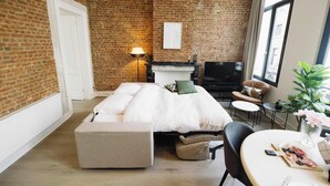 A modern room with a brick wall, a bed with white bedding, a TV, a small table with food, and a plant. - Bright 1-Bedroom Apartment with Balcony in Antwerp (Antwerpen)
