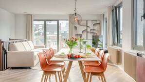 Ein stilvoller Essbereich mit Tisch für vier Personen, umgeben von orangefarbenen Stühlen, einer Couch und einem großen Fenster mit Blick auf die Stadt.
