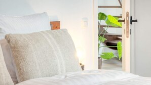 Ein Schlafzimmer mit einem Bett, Kissen, einem Nachttisch, einer Tür zu einer Treppe und einer Pflanze.