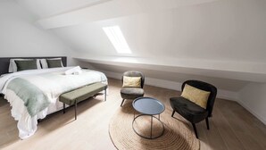 Quarto com cama, um banquinho, duas cadeiras, uma mesinha e um claraboia.