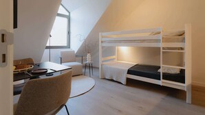 Una habitación moderna y compacta con literas, una zona de comedor con mesa y sillas, y una mesita auxiliar con un jarrón.