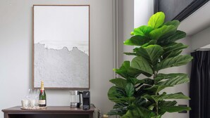 Una stanza con un quadro astratto incorniciato, un tavolo con una bottiglia di champagne, bicchieri, una macchina per il caffè e una grande pianta verde.