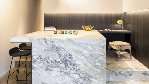 A modern kitchen with a marble countertop, two bar stools, a basket of fruit, and a small table with a lamp. - Secret & private suite with timeless elegance (Antwerpen)