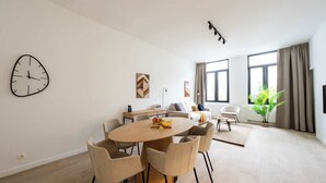 Un saló modern amb una taula rodona de fusta per dinar a la qual s'ajunten sis cadires, un sofà i una taula baixa amb fruita. Hi ha un rellotge de paret gran, dos quadres abstractes i una planta en test.