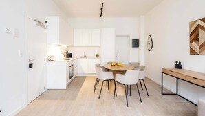 Una cuina moderna amb armaris blancs, una zona de menjador amb taula rodona i cadires, i un petit escriptori amb cadira.