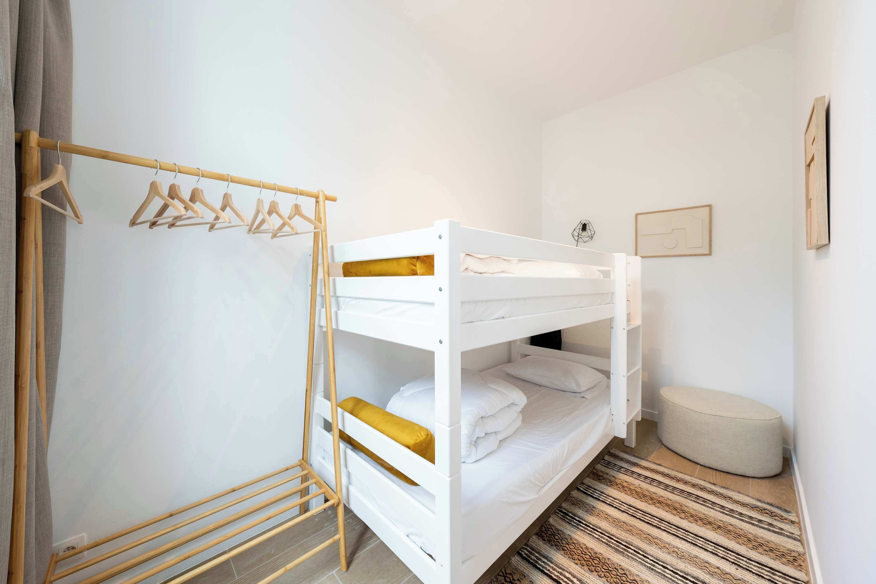 Un lit superposé avec une échelle en bois, un portant à vêtements suspendu, un petit pouf et un tapis à motifs.