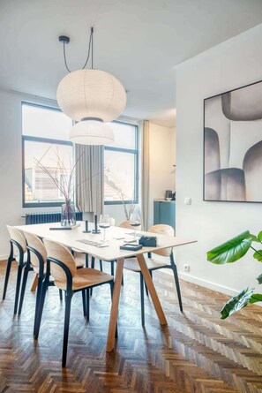 Ein moderner Essbereich mit Holzboden im Fischgrätmuster, einem runden Tisch mit Stühlen, einer Pendelleuchte und einem großen Fenster mit Vorhängen.