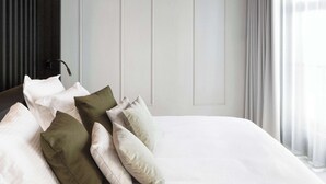 Un lit bien fait, avec du linge de lit blanc et plusieurs oreillers, dont un vert, dans une pièce dont un mur est recouvert de lambris en bois foncé et qui possède une porte blanche.