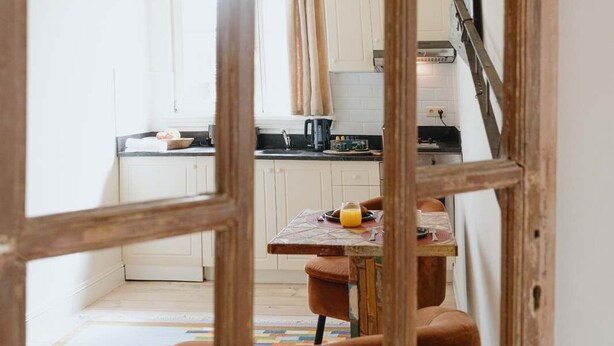 A view through a wooden-framed window into a kitchen with white cabinets, a countertop, and a table with a colorful rug. - Cozy Hideaway Near the Heart of Antwerp (Antwerpen)