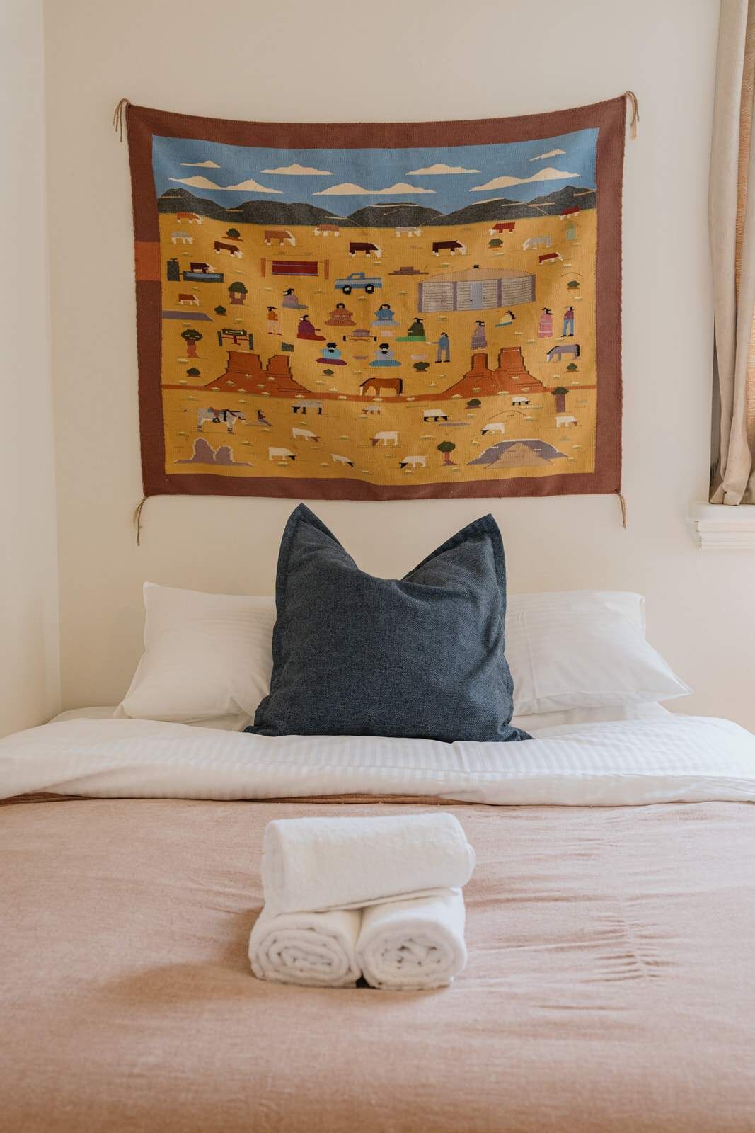 Un letto rifatto con lenzuola bianche e un asciugamano piegato, un cuscino grigio scuro e una tappezzeria che raffigura una scena campestre sopra il letto.