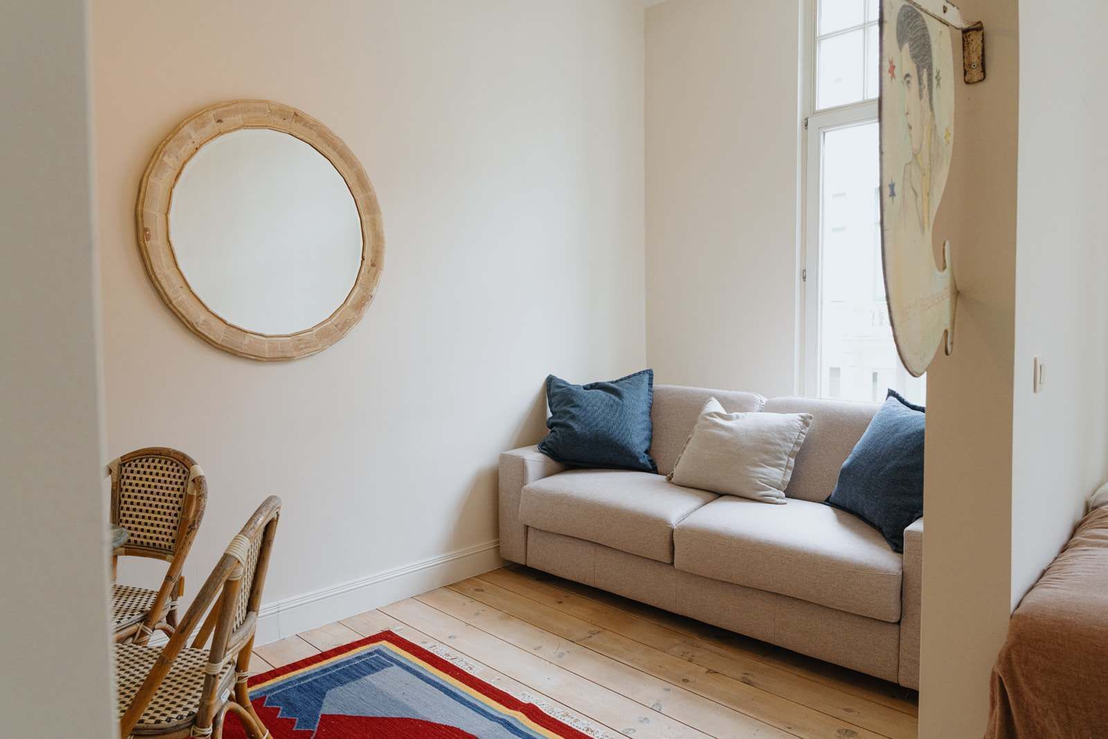 A room with a beige sofa, a round mirror, a wicker chair, and a colorful rug.