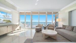 Amplia sala de estar con un ventanal que da a una playa y al océano. Hay un sofá, dos sillones, una mesa de centro y un televisor montado en la pared.