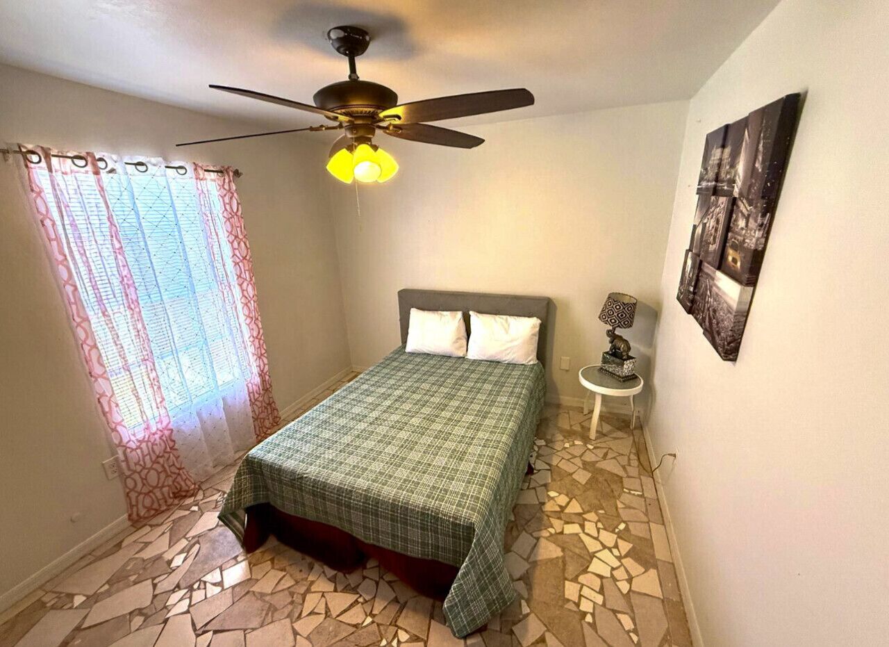 A bedroom with a bed, a ceiling fan, a small table with a lamp, and a wall-mounted picture.