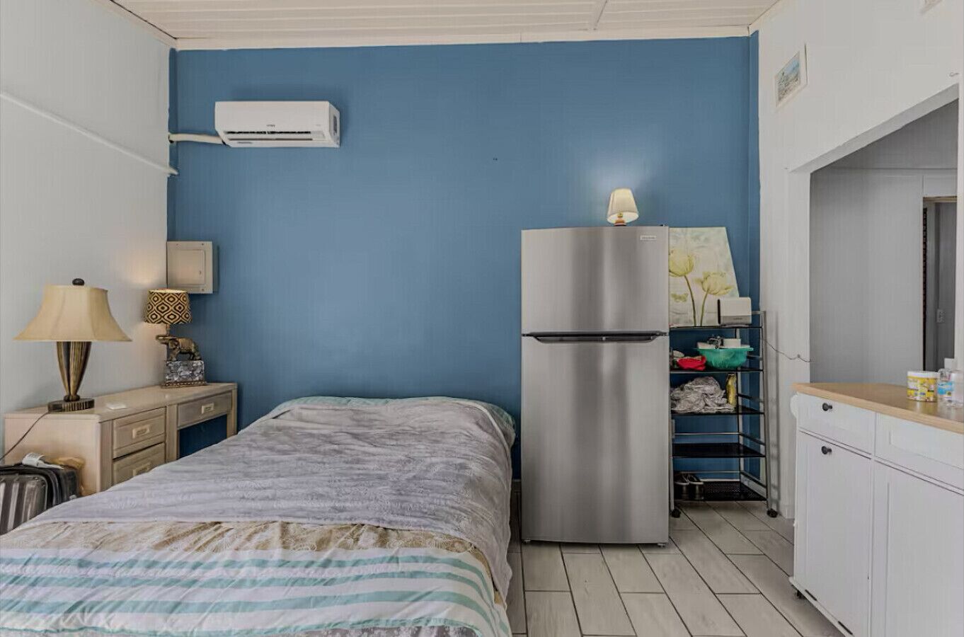 A bedroom with a bed, a dresser, a refrigerator, a lamp, and a wall-mounted air conditioner.