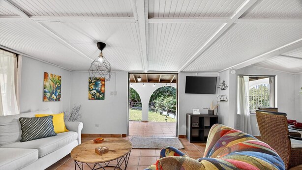 A spacious living area with a white ceiling, a sofa with patterned cushions, a colorful armchair, a wooden coffee table, and a view of a garden through an open doorway.