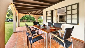 An outdoor patio with a wooden table and chairs, a tiled floor, and a view of a garden and parked cars.