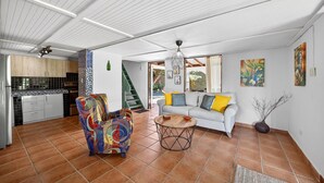 A spacious living area with a sofa, colorful chair, and a round coffee table. A kitchen with wooden cabinets and a tiled backsplash is visible.