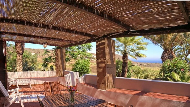 A covered outdoor seating area with a table and chairs, a view of the landscape, and a clear sky. - Dammuso Ibiscus mit Blick aufs Wasser by Interhome (Pantelleria)