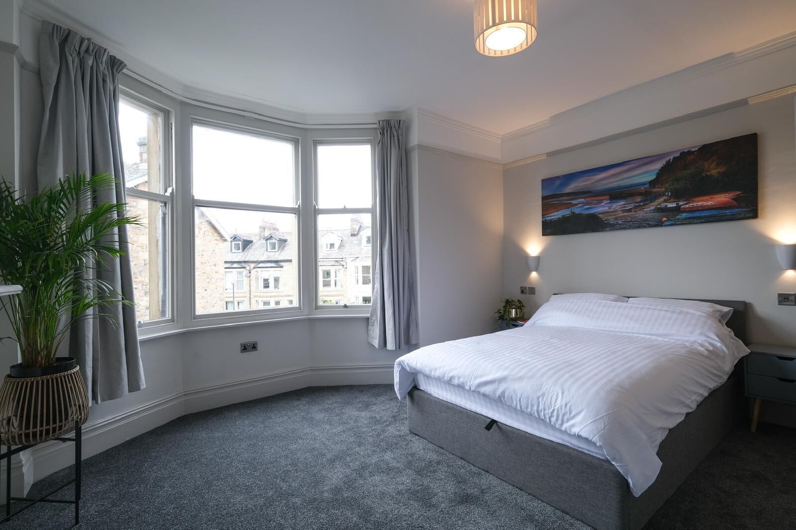 A bedroom with a large bed, a potted plant, a painting, and a view of buildings through large windows.