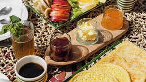 Uma mesa de café da manhã com torradas, frutas, chá e acompanhamentos sobre um tapete tecido.